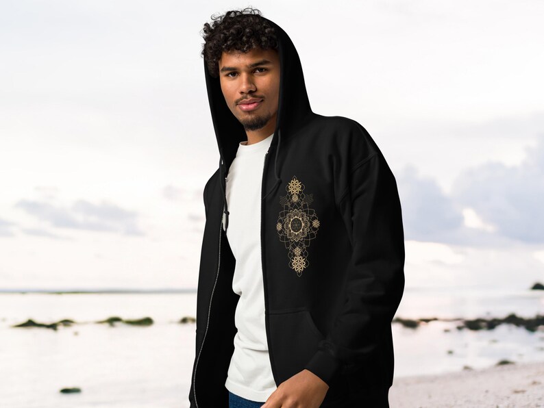 May include: Black zip-up hoodie with a gold geometric design on the front. The hoodie has a hood and a white t-shirt underneath. The person is standing in front of a beach.