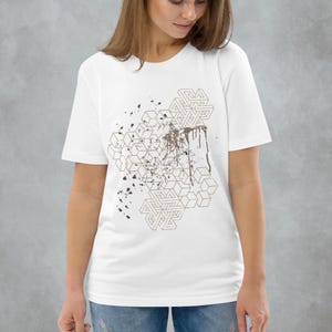 Puede incluir: Camiseta blanca con un diseño geométrico con detalles marrones y salpicaduras. El diseño incluye formas entrelazadas y un efecto de goteo, creando un aspecto moderno y artístico. La camiseta es de material suave.