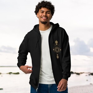 May include: Black zip-up hoodie with a gold geometric design on the chest. The hoodie has a drawstring hood and a full-length zipper. The person is wearing a white t-shirt underneath. The background is a beach scene.