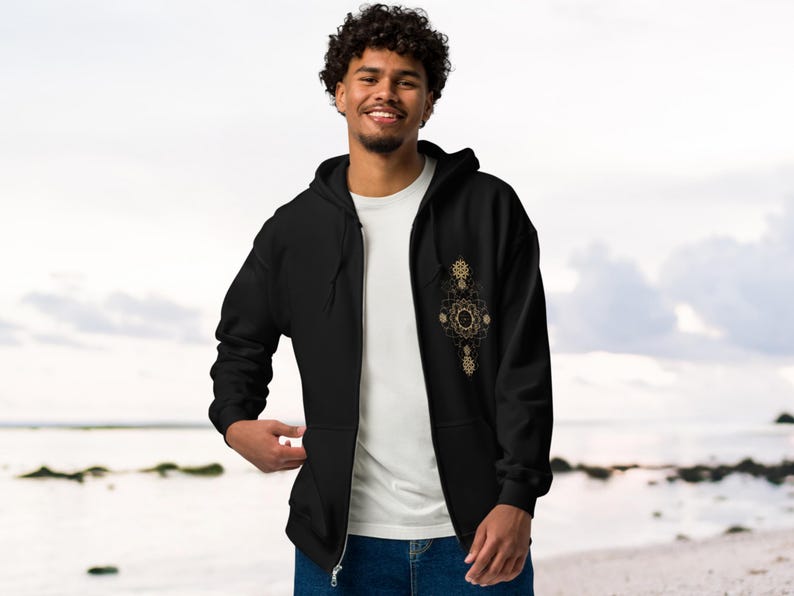May include: Black zip-up hoodie with a gold geometric design on the chest. The hoodie has a drawstring hood and a full-length zipper. The person is wearing a white t-shirt underneath. The background is a beach scene.