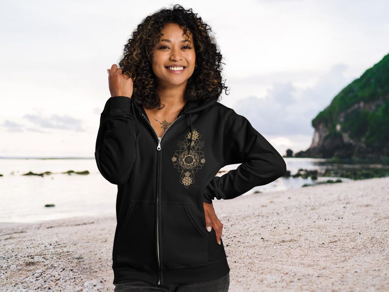 May include: Black zip-up hoodie with a gold geometric design on the front. The hoodie has a full zipper and a hood. The design is made up of circles and lines. The person is standing on a beach.