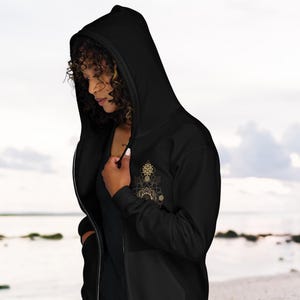 May include: Black zip-up hoodie with a hood and a gold-colored geometric design on the chest. The hoodie has a front pocket and a zipper closure. The person in the image is wearing the hoodie.
