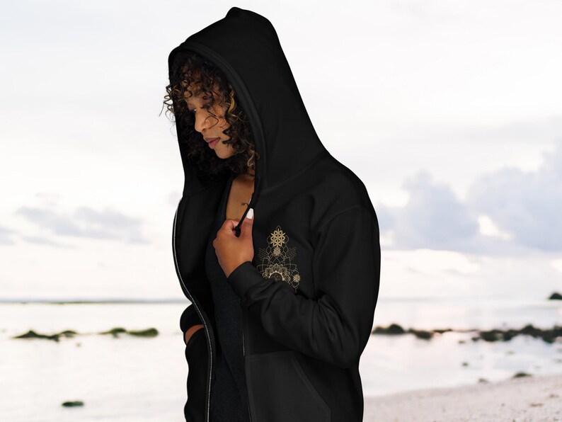May include: Black zip-up hoodie with a hood and a gold-colored geometric design on the chest. The hoodie has a front pocket and a zipper closure. The person in the image is wearing the hoodie.
