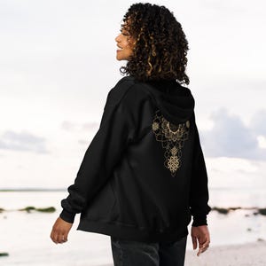 May include: Black zip-up hoodie with a gold mandala design on the back. The intricate pattern features floral and geometric shapes. The hoodie has a hood and is made of a soft material.