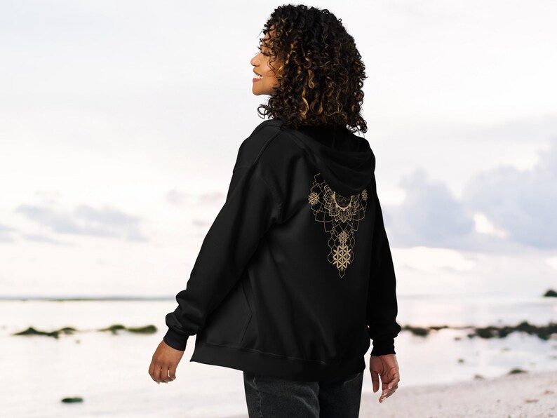 May include: Black zip-up hoodie with a gold mandala design on the back. The intricate pattern features floral and geometric shapes. The hoodie has a hood and is made of a soft material.