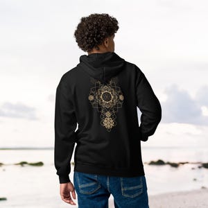 May include: Black hoodie with a gold geometric design on the back. The design features a central circular element with radiating patterns and a series of interconnected shapes. The hoodie has a hood and long sleeves.
