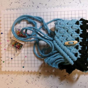 May include: A blue and black macrame wall hanging with fringe, pinned to a white gridded foam board. A small container of colorful pins is also visible.
