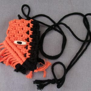 Puede incluir: Una pequeña bolsa de crochet naranja y negra con un detalle de ojo blanco y un cordón negro.