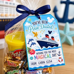 May include: A clear cellophane bag tied with a navy blue ribbon, containing a yellow Play-Doh container and assorted mini candy bars. A blue tag reads "You've Been Pixie Dusted!" with a cruise ship illustration and the text "Wishing you the most Magical Day!"