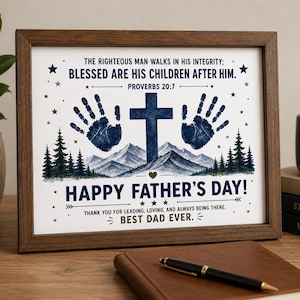 May include: Framed print with a brown wooden frame. The print features a blue cross, handprints, and mountain scenery. Text includes "Happy Father's Day!" and a Bible verse. A pen and a leather-bound notebook are in the foreground.