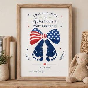May include: A framed print featuring the text I WAS THIS LITTLE ON America's 250th BIRTHDAY, a red, white, and blue bow, and two dark blue baby footprints. Below are a small red heart, the date JULY 4, 2026, and the text made with love by.