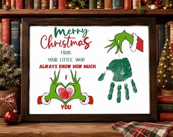 Op de afbeelding: Een ingelijste print met de tekst Merry Christmas FROM: YOUR LITTLE WHO ALWAYS KNOW HOW MUCH I YOU. Het ontwerp bevat groene cartoonhanden die een hart vormen rond een rode vingerafdruk en een groene handafdruk onder een groene mouw.