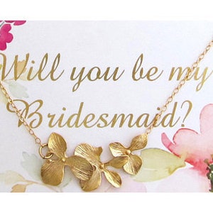 May include: A white card with watercolor flowers and the text "Will you be my bridesmaid?" A gold necklace with a floral pendant hangs from the card.