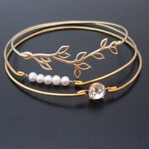 Pode incluir: Tr&ecirc;s pulseiras de bracelete douradas com um delicado design de ramo e um &uacute;nico charme de strass. A pulseira do meio tem uma fila de cinco p&eacute;rolas brancas.