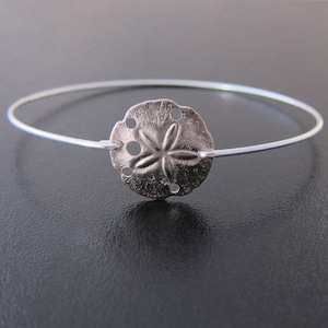 May include: A silver bangle bracelet with a sand dollar charm. The sand dollar charm has a five-pointed star design.