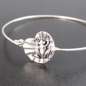 May include: A silver bangle bracelet with a silver frog charm. The frog is sitting on a lily pad.