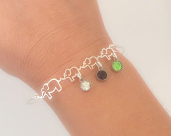 May include: A silver bracelet with a chain of elephant cutouts. The bracelet has three charms hanging from it: a clear crystal, a black crystal, and a green crystal.