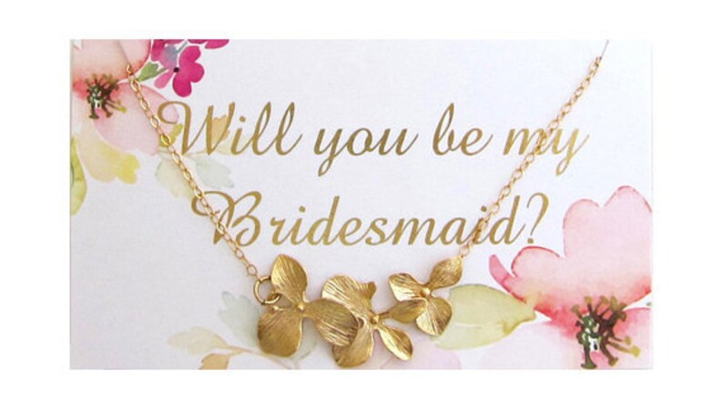 May include: A white card with watercolor flowers and the text "Will you be my bridesmaid?" A gold necklace with a floral pendant hangs from the card.