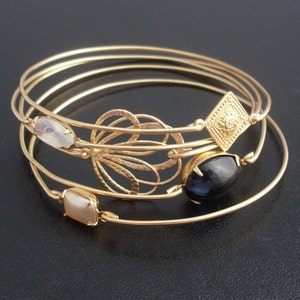 May include: Four gold bangle bracelets with different charms. The charms include a black oval stone, a white rectangular stone, a gold flower design, and a gold square with a design.