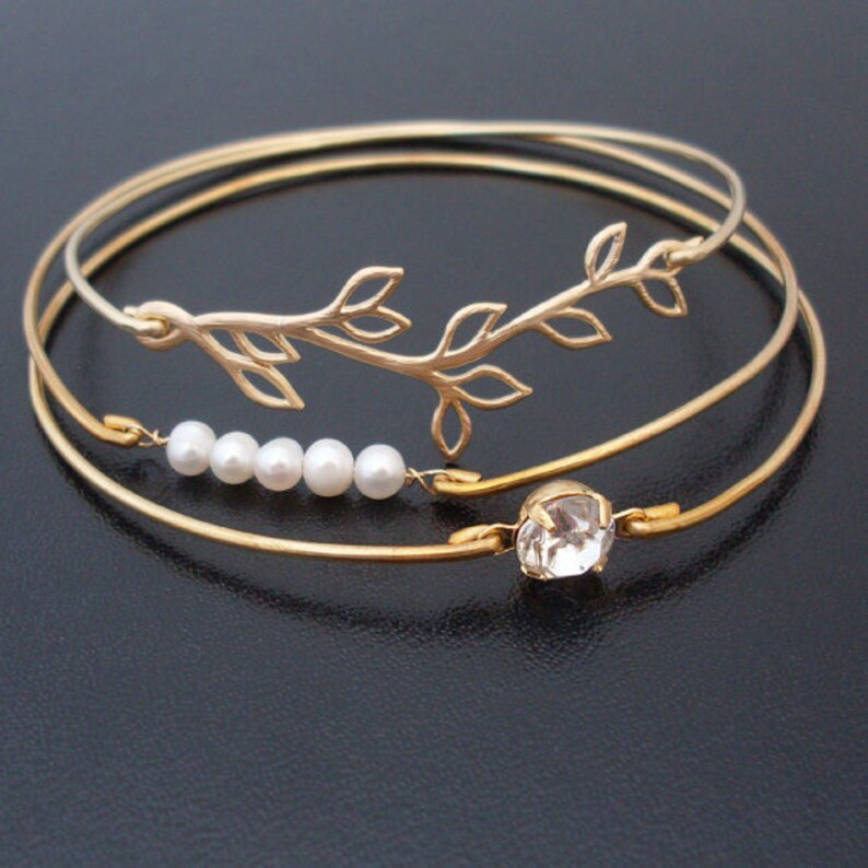 Pode incluir: Tr&ecirc;s pulseiras de bracelete douradas com um delicado design de ramo e um &uacute;nico charme de strass. A pulseira do meio tem uma fila de cinco p&eacute;rolas brancas.