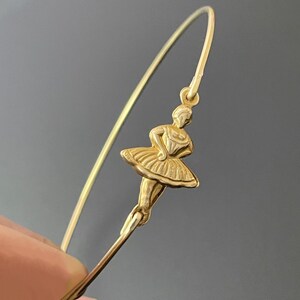 May include: A gold-toned bangle bracelet with a delicate ballerina charm. The ballerina is in a dancing pose with her arms outstretched.
