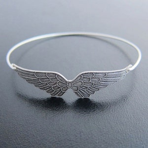 K&ouml;nnte beinhalten: Ein silbernes Armreif-Armband mit einem detaillierten Engelsfl&uuml;gel-Design.