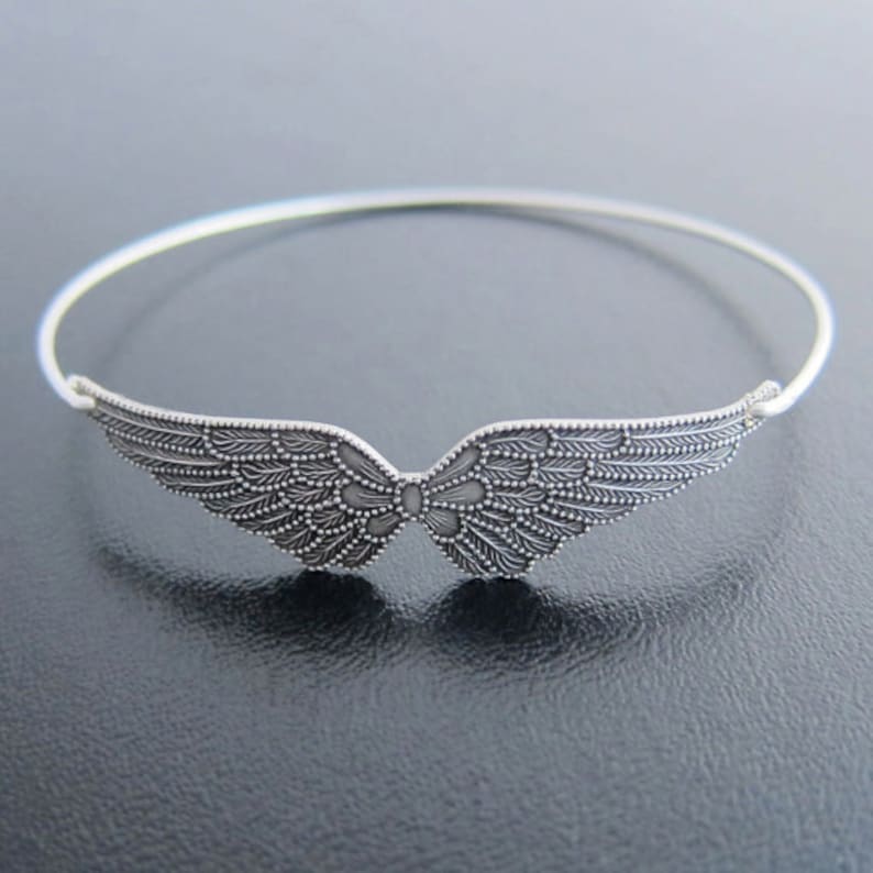 K&ouml;nnte beinhalten: Ein silbernes Armreif-Armband mit einem detaillierten Engelsfl&uuml;gel-Design.