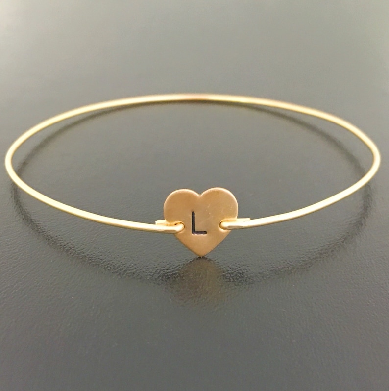 Puede incluir: Un brazalete dorado con un dije en forma de coraz&oacute;n con la letra "L" grabada.