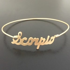 May include: Gold bangle bracelet with the word "Scorpio" written in a cursive font.