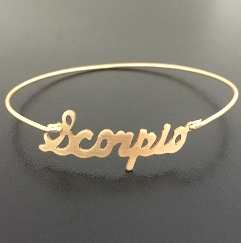 May include: Gold bangle bracelet with the word "Scorpio" written in a cursive font.