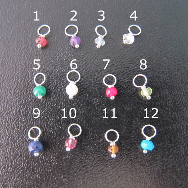 May include: Twelve silver jump rings with different colored gemstones attached. The jump rings are numbered 1 through 12. The gemstones are red, purple, white, clear, green, white, red, green, blue, red, brown, and blue.
