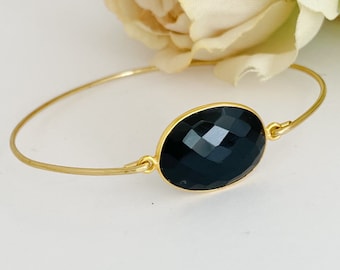 May include: A gold bangle bracelet with a large, oval-shaped black gemstone. The gemstone is faceted and has a high polish.
