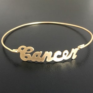 May include: Gold bangle bracelet with the word "Cancer" written in a cursive script.