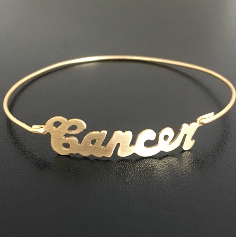 May include: Gold bangle bracelet with the word "Cancer" written in a cursive script.