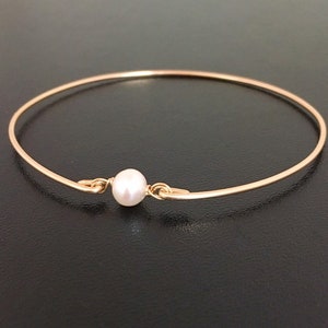 May include: A delicate gold-tone bangle bracelet featuring a single, lustrous white pearl at the center. The bracelet has a simple, elegant design, perfect for everyday wear. The pearl is approximately 0.3 inches in diameter.