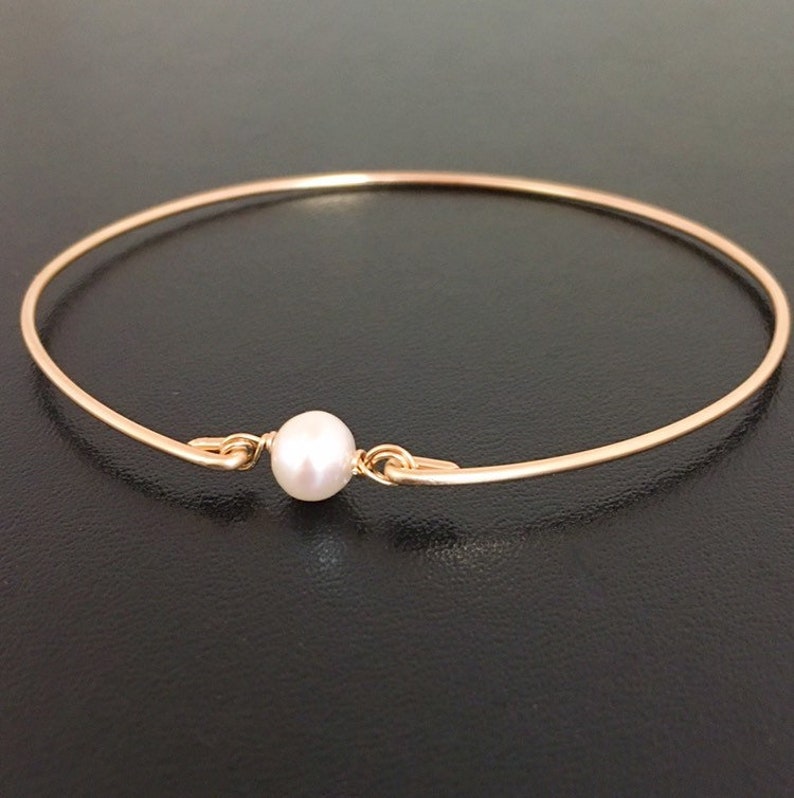 May include: A delicate gold-tone bangle bracelet featuring a single, lustrous white pearl at the center. The bracelet has a simple, elegant design, perfect for everyday wear. The pearl is approximately 0.3 inches in diameter.