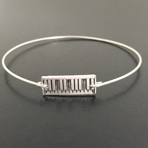 May include: A silver bangle bracelet with a piano key charm. The charm is a rectangular shape with a detailed design of piano keys.