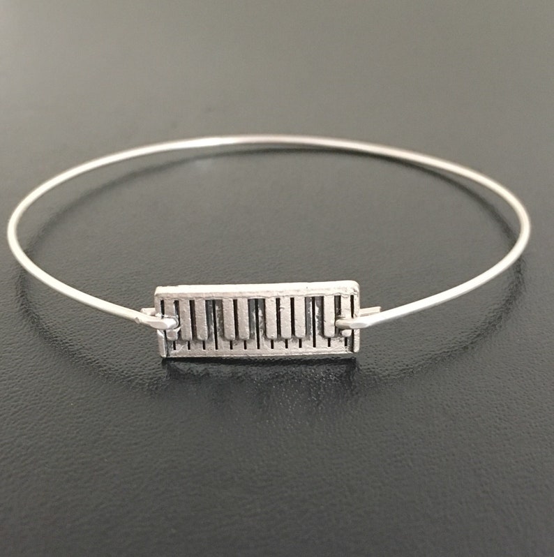 May include: A silver bangle bracelet with a piano key charm. The charm is a rectangular shape with a detailed design of piano keys.
