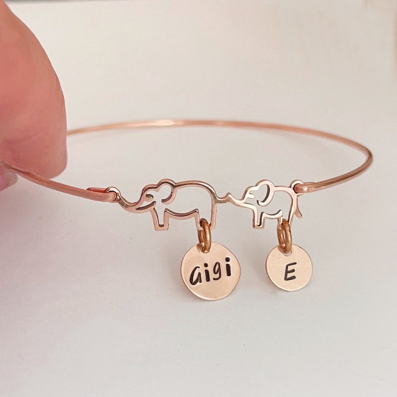 May include: Rose gold bangle bracelet with two elephant charms and personalized discs. The larger disc says "gigi" and the smaller disc says "E".
