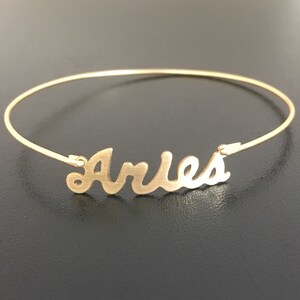 May include: A gold-toned bangle bracelet with the word "Aries" written in a cursive font.