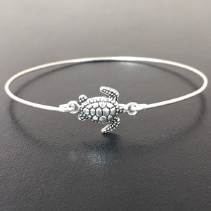 May include: A silver bangle bracelet with a small turtle charm. The turtle is facing to the right and has a detailed shell.