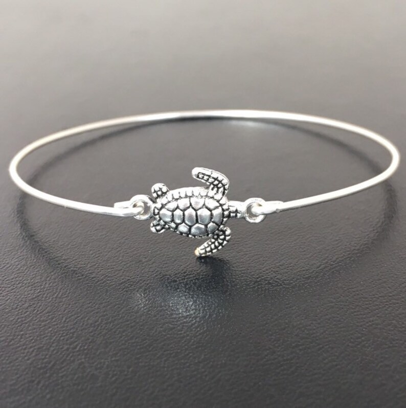 May include: A silver bangle bracelet with a small turtle charm. The turtle is facing to the right and has a detailed shell.
