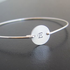 Op de afbeelding: Een zilveren armband met een ronde bedel met de letter "E" erop gegraveerd.
