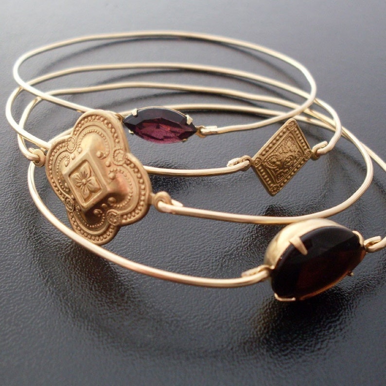 May include: A set of four gold-toned bangles. Three bangles feature decorative elements: a quatrefoil, a diamond shape, and a marquise-cut dark purple stone. The bangles are displayed on a dark surface.