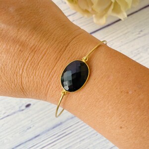 May include: A gold bangle bracelet with a black oval gemstone. The bracelet is on a person's wrist.