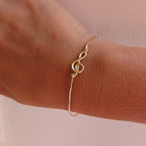 May include: A gold-tone bracelet with a treble clef charm. The bracelet is on a person's wrist.