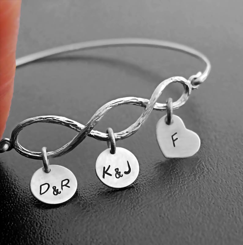 May include: Silver infinity bracelet with three charms: a heart charm engraved with the letter "F", a round charm engraved with "K&J", and a round charm engraved with "D&R".