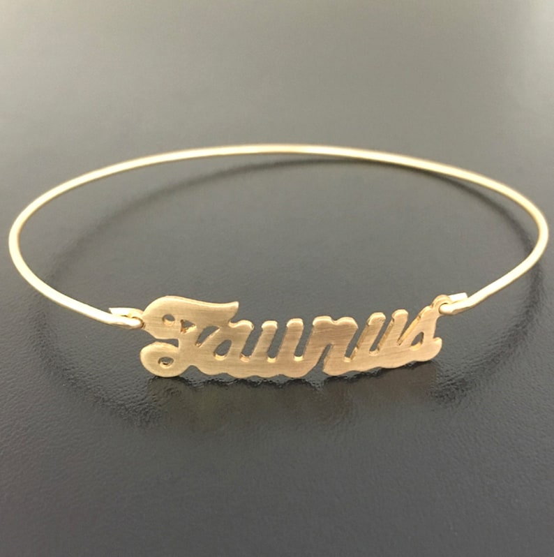 May include: A gold-toned bangle bracelet with the word "Taurus" written in a cursive font.