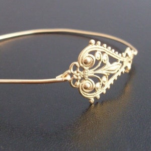 May include: A gold-colored bracelet with an ornate filigree design. The decorative element features a symmetrical pattern of swirls, leaves, and small circular details. The bracelet is set against a dark background, highlighting its intricate craftsmanship.