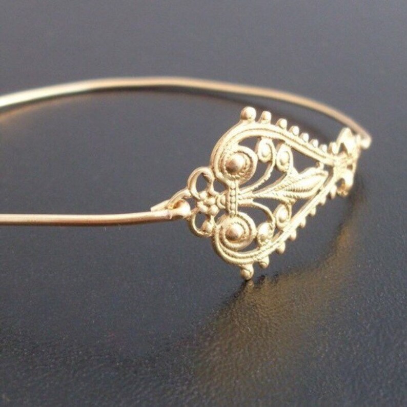 May include: A gold-colored bracelet with an ornate filigree design. The decorative element features a symmetrical pattern of swirls, leaves, and small circular details. The bracelet is set against a dark background, highlighting its intricate craftsmanship.